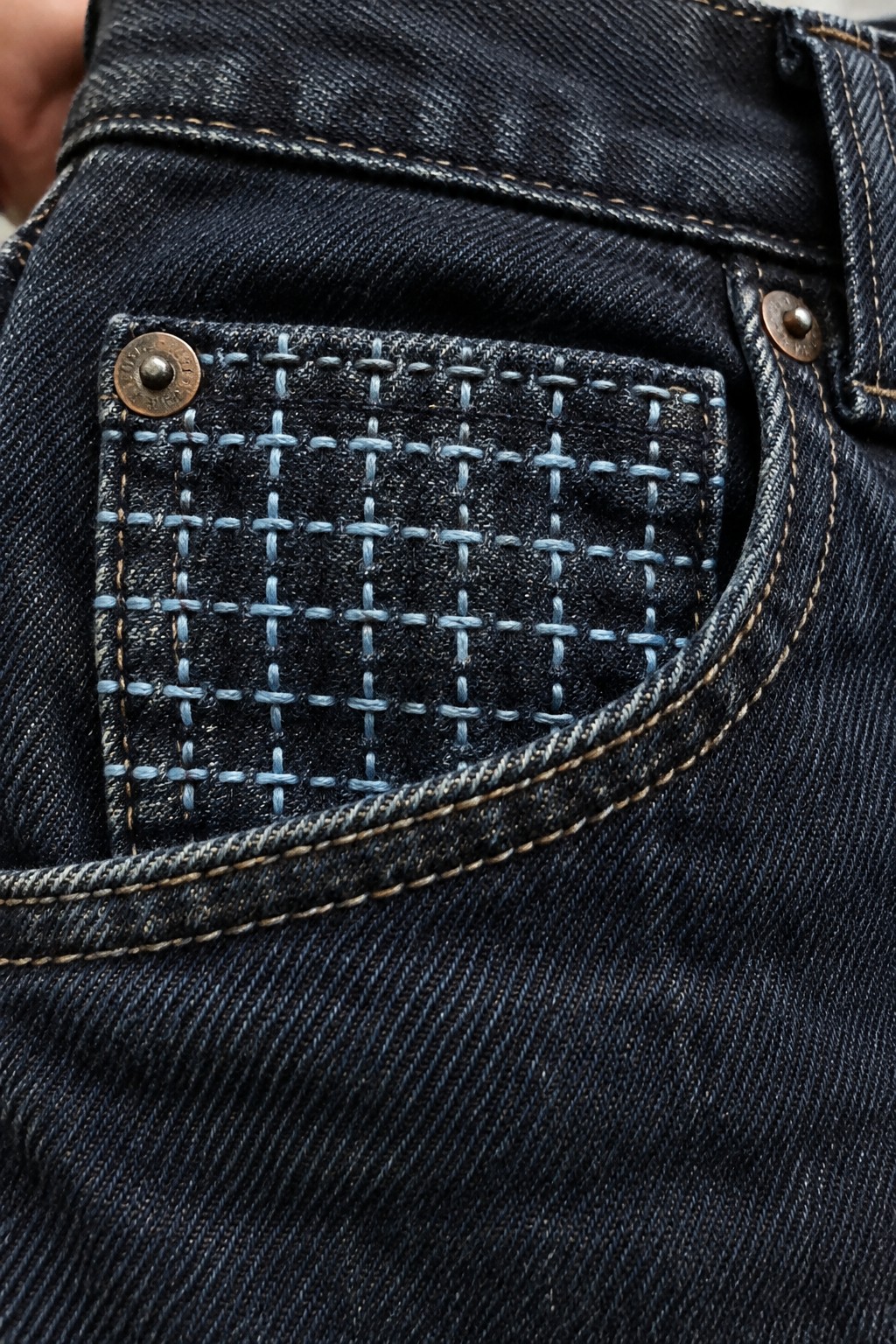 Pale blue grid stitching covers a dark denim jeans pocket with copper-colored rivets.