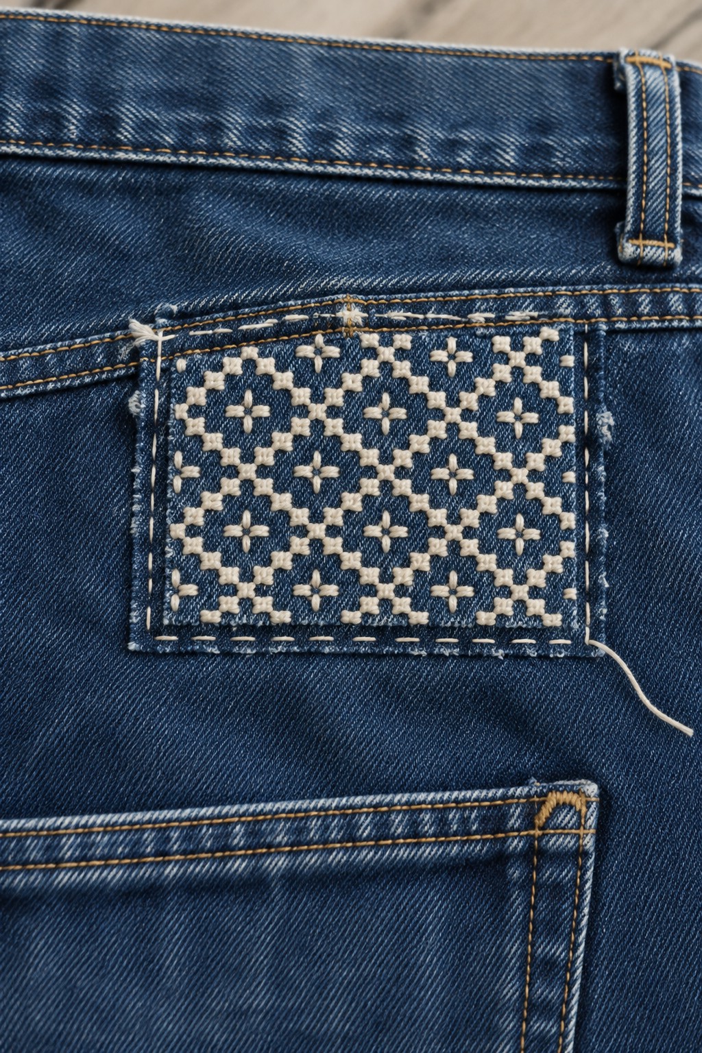 Cream geometric sashiko stitching forms a diamond lattice on a rectangular denim patch sewn over a jeans pocket.