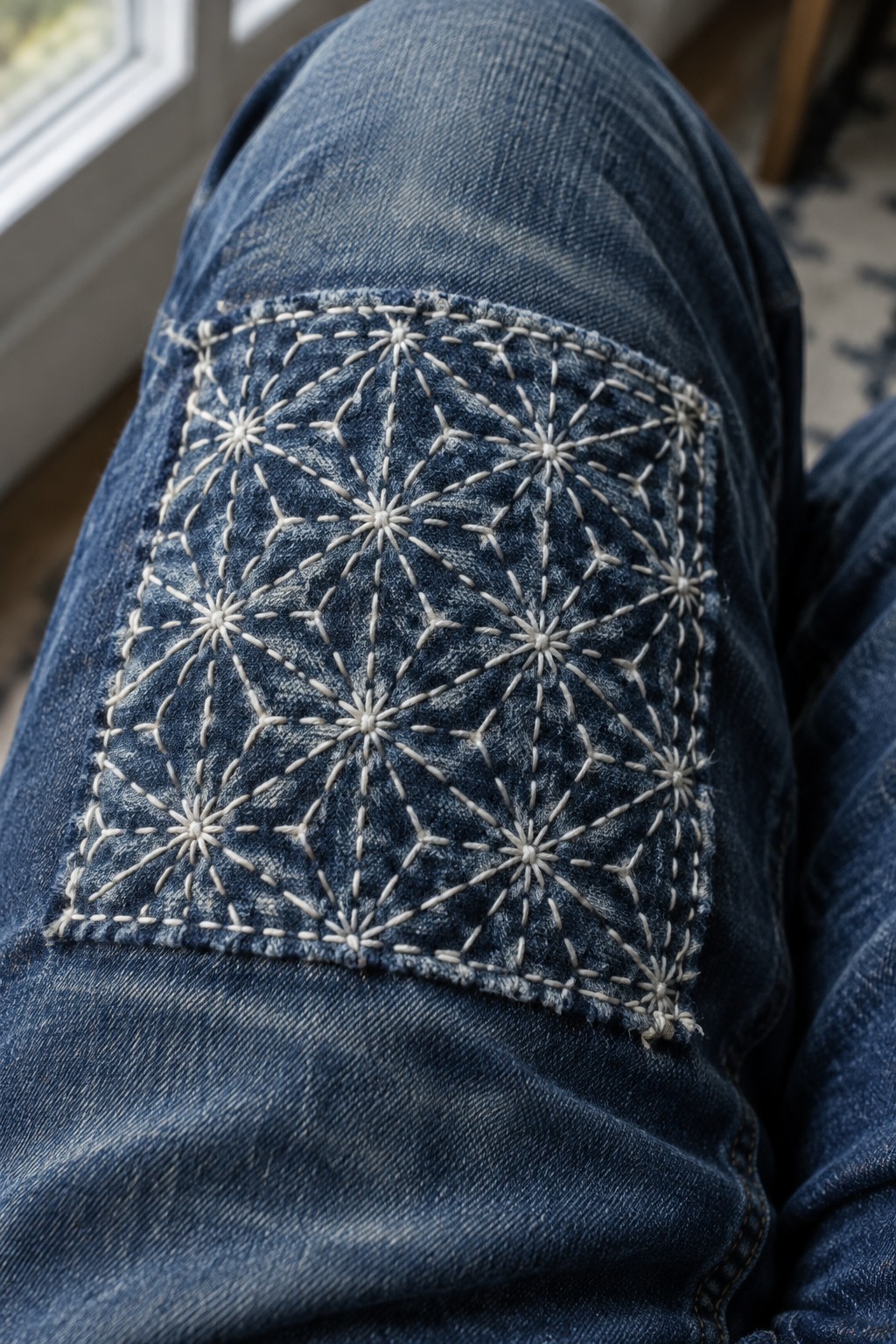 Pale starburst stitching and connecting lines cover a rectangular patch on the knee of blue jeans.