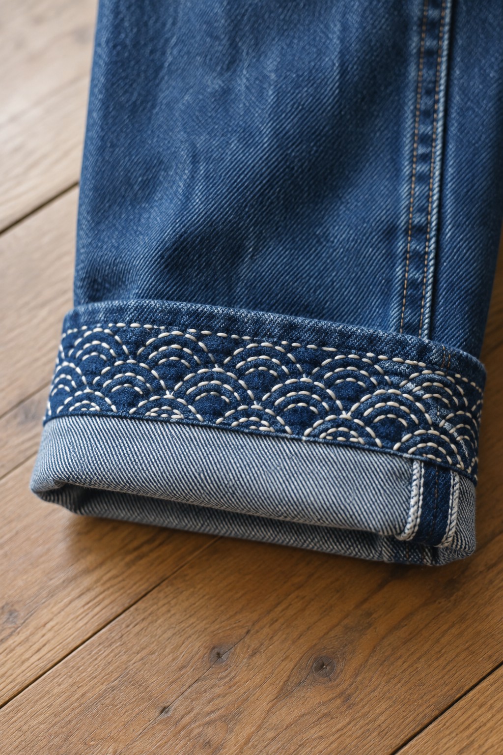 Close-up of a dark denim jeans cuff embroidered with rows of pale scalloped stitching.