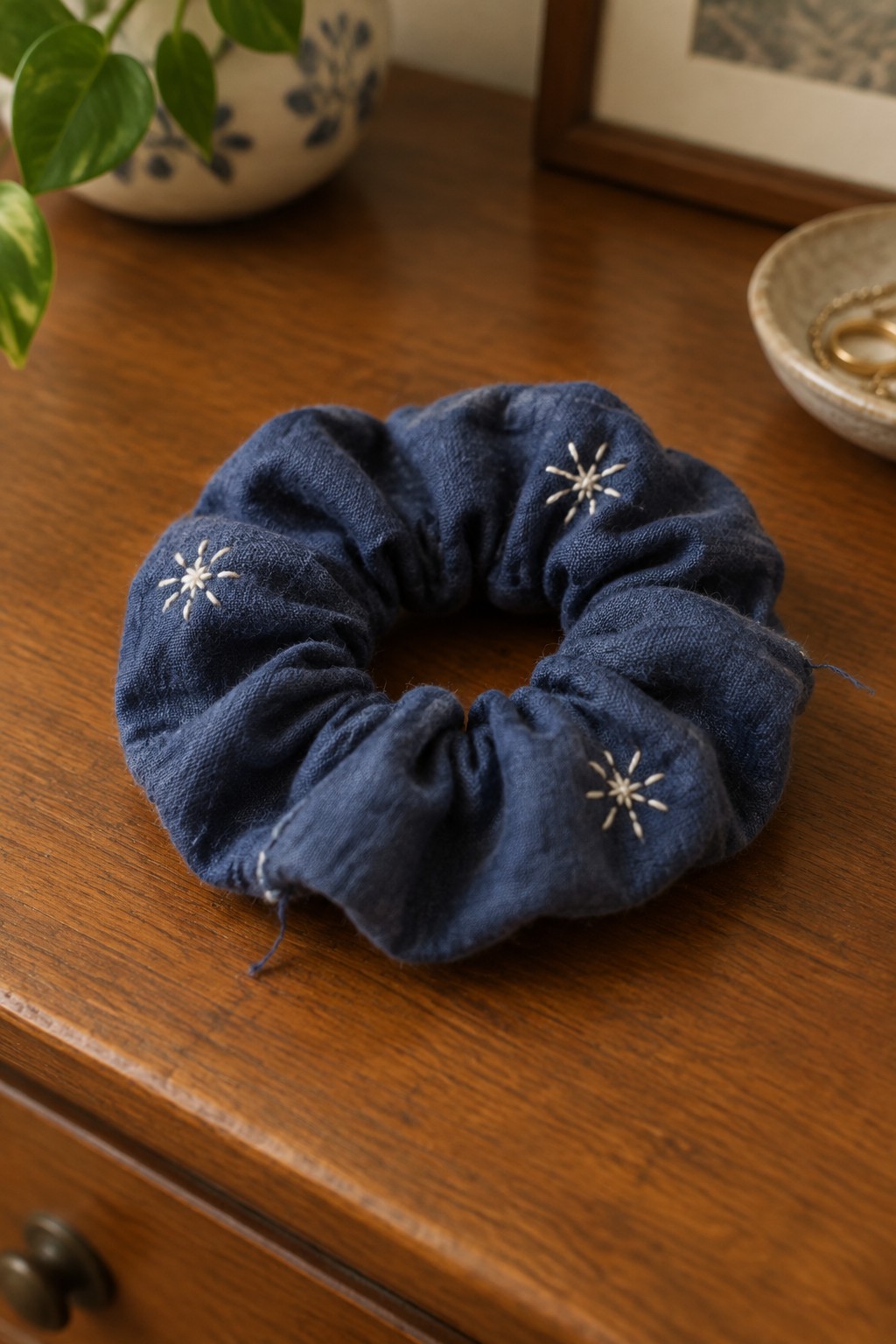 A navy gathered fabric scrunchie with small cream starburst embroidery rests on a wooden dresser.