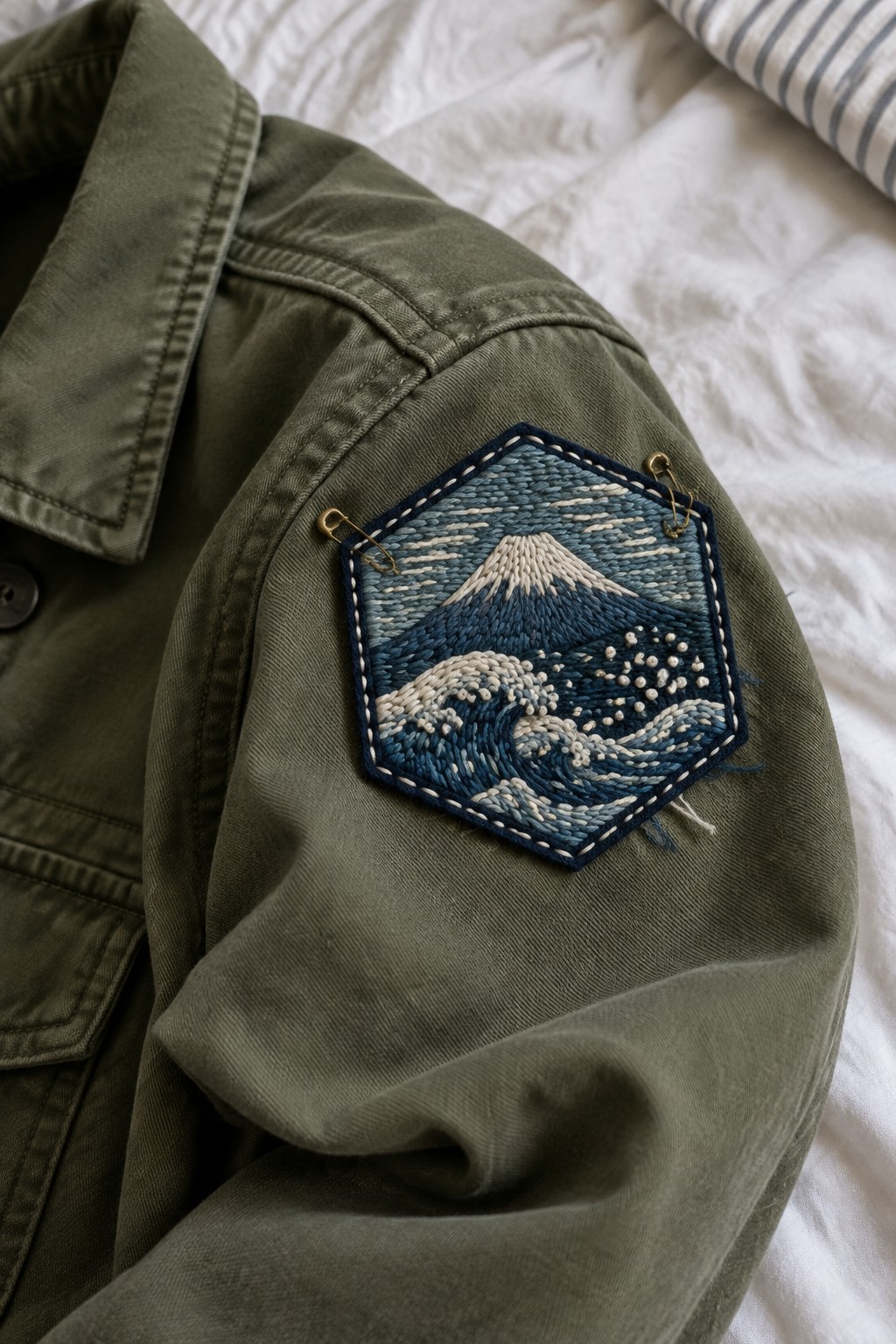 A blue-and-cream embroidered hexagonal patch with a mountain and waves is pinned to the sleeve of an olive jacket.