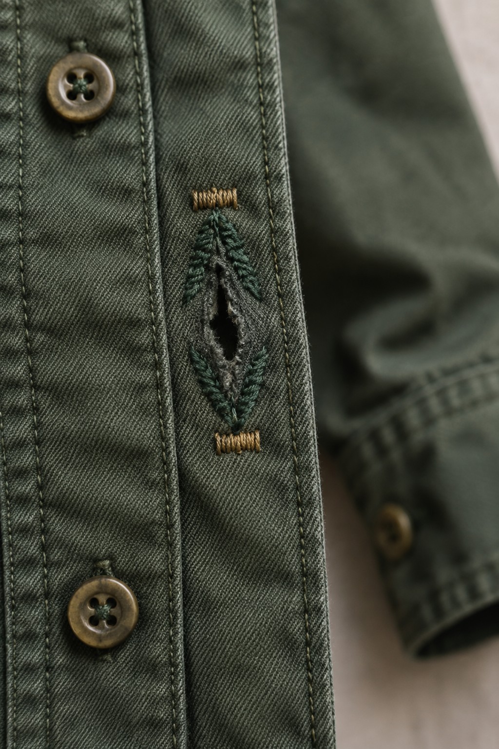 Close-up of a green shirt placket with a buttonhole reinforced by teal and tan embroidery beside two brown buttons.