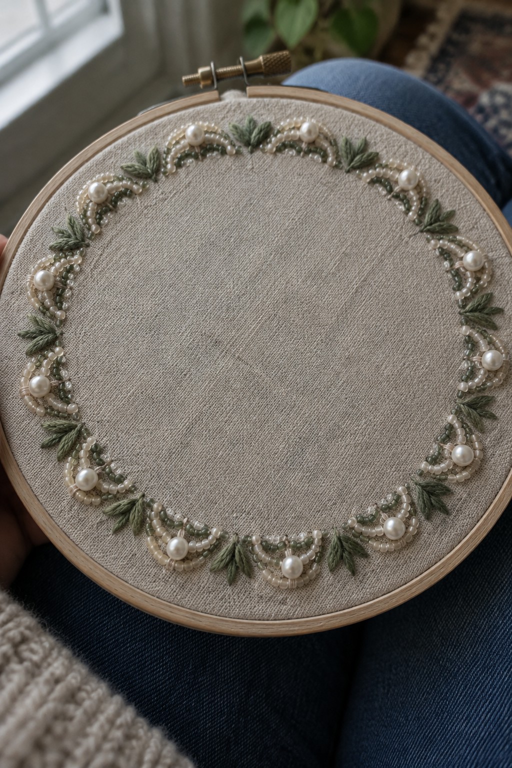 A fabric embroidery hoop with a circular border of pale beads, pearl-like accents, and green leaf motifs.