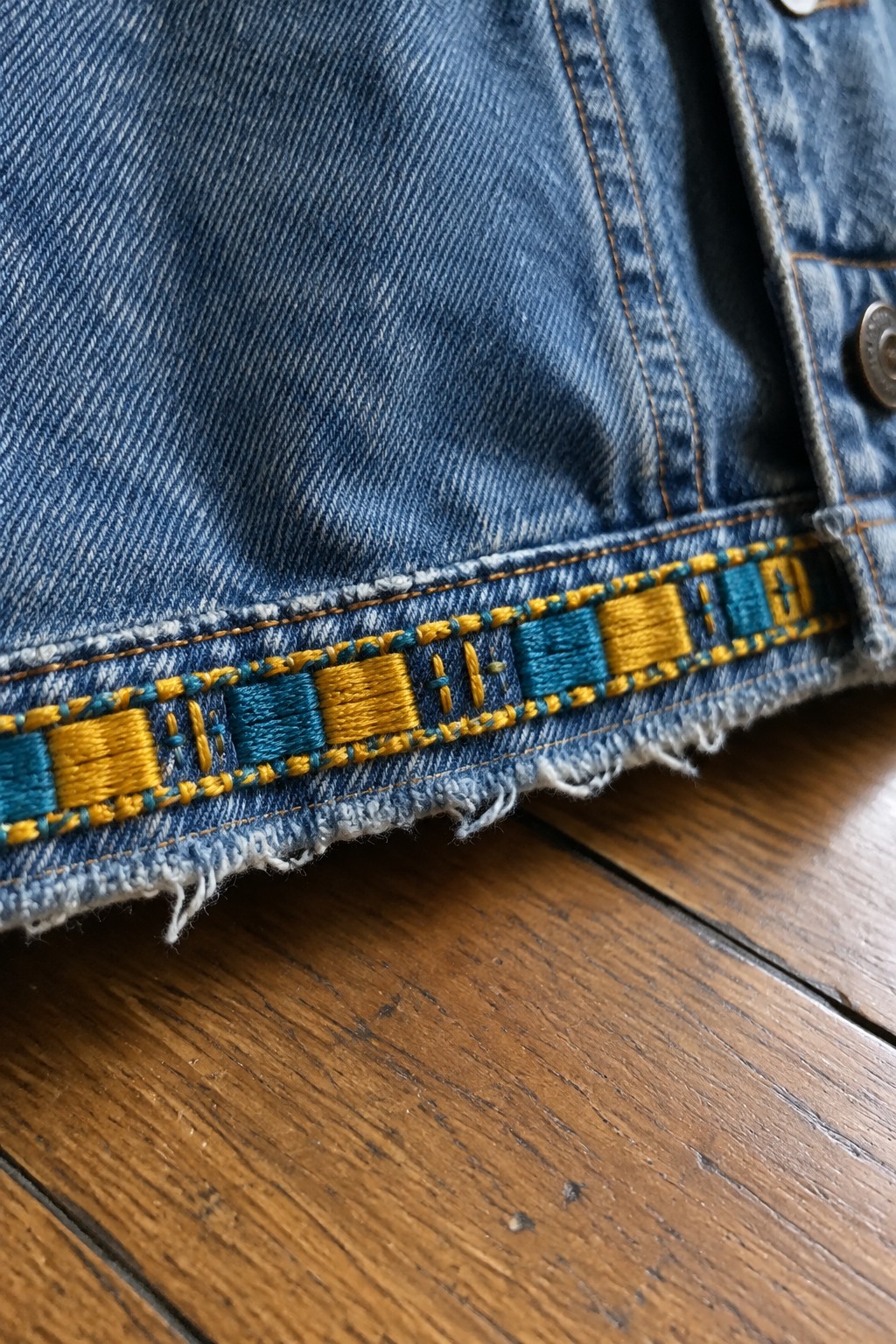 Teal and golden geometric embroidery runs along the frayed hem of a blue denim jacket.