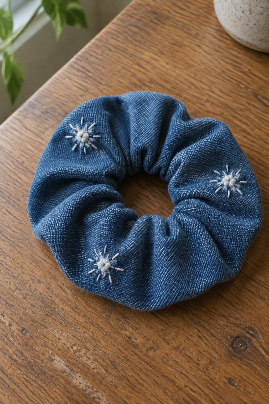 A blue denim scrunchie with three white embroidered snowflakes rests on a wooden table.