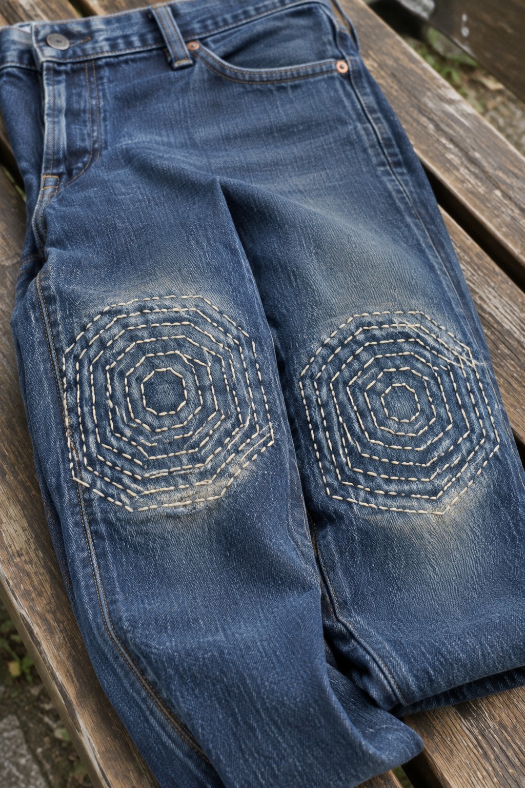 Blue jeans with pale concentric geometric stitching across both knees, laid on a wooden bench.