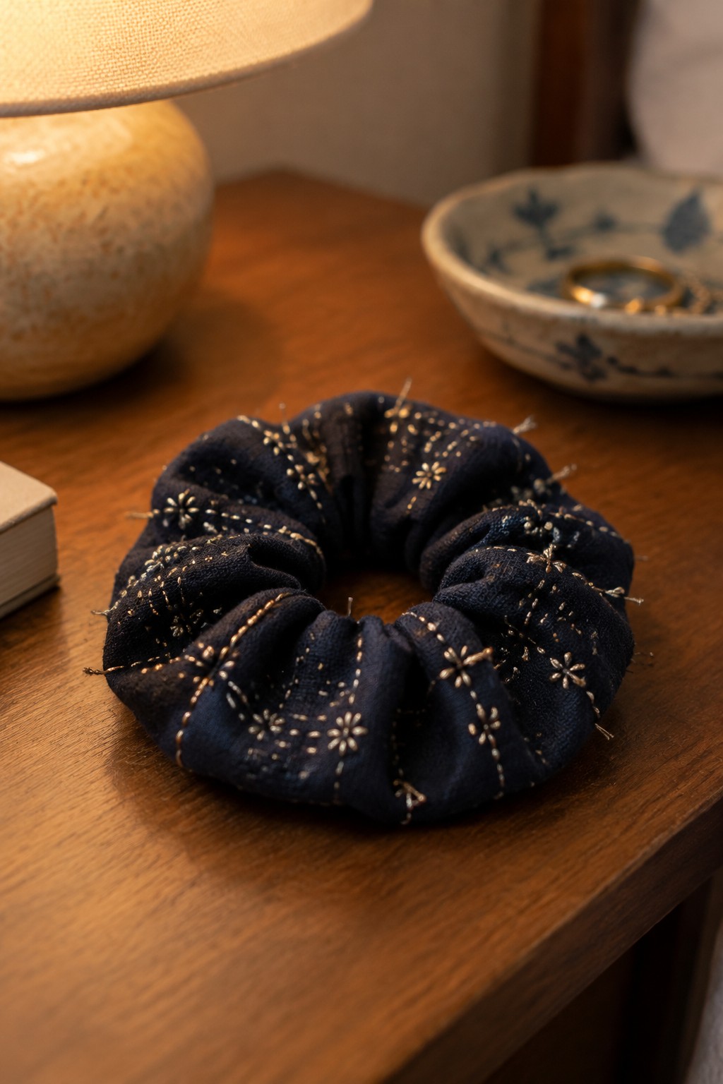 A dark gathered fabric scrunchie with small light-colored embroidered motifs rests on a wooden table.