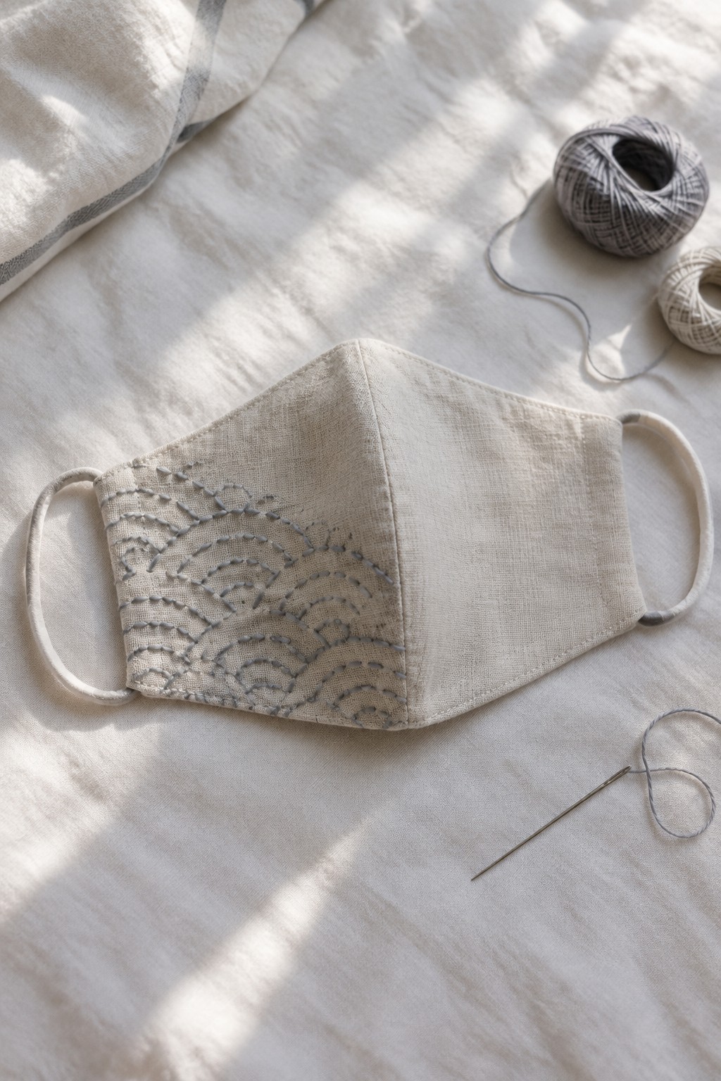 A light fabric face mask with gray wave embroidery lies on linen beside thread and a needle.