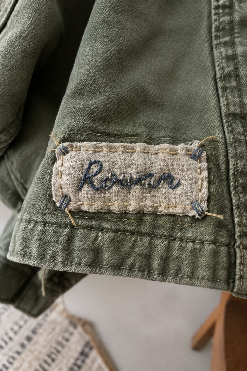 A pale rectangular patch with a blue embroidered name is stitched onto the cuff of an olive-green jacket.