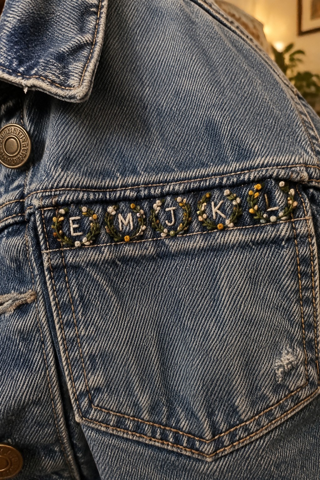 A denim jacket pocket embroidered with cream letters surrounded by green leaves and small yellow and white details.