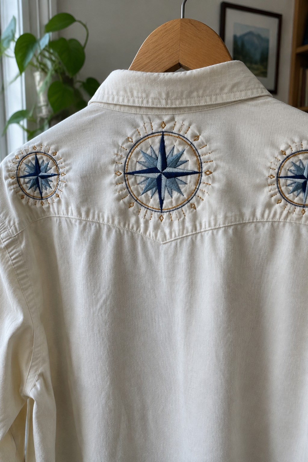 A white shirt on a wooden hanger embroidered with blue compass rose circles and gold accents across the back yoke.