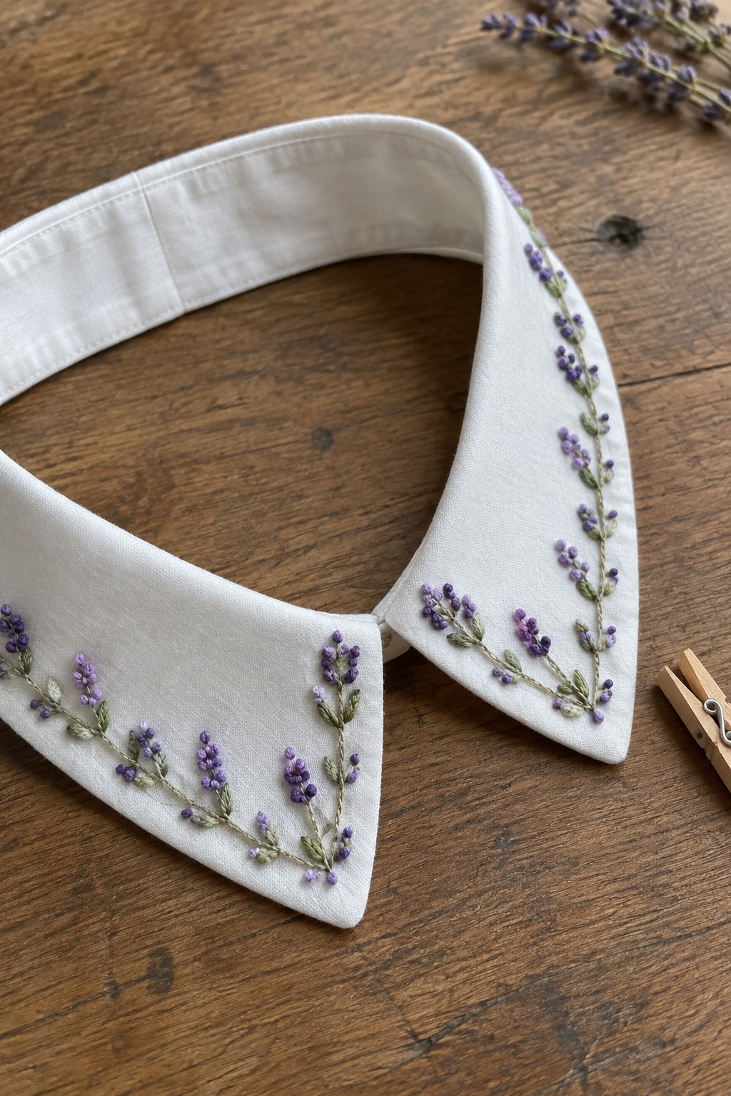 A white pointed collar embroidered with purple flowers and green stems lies on a wooden table.