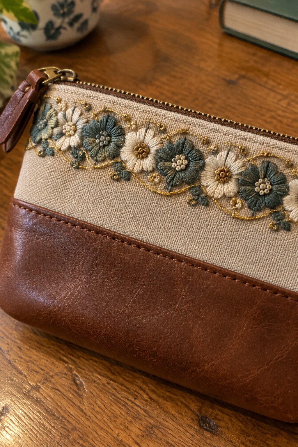 A brown zippered pouch with a beige panel embroidered with a repeating border of blue-gray and cream flowers with gold accents.
