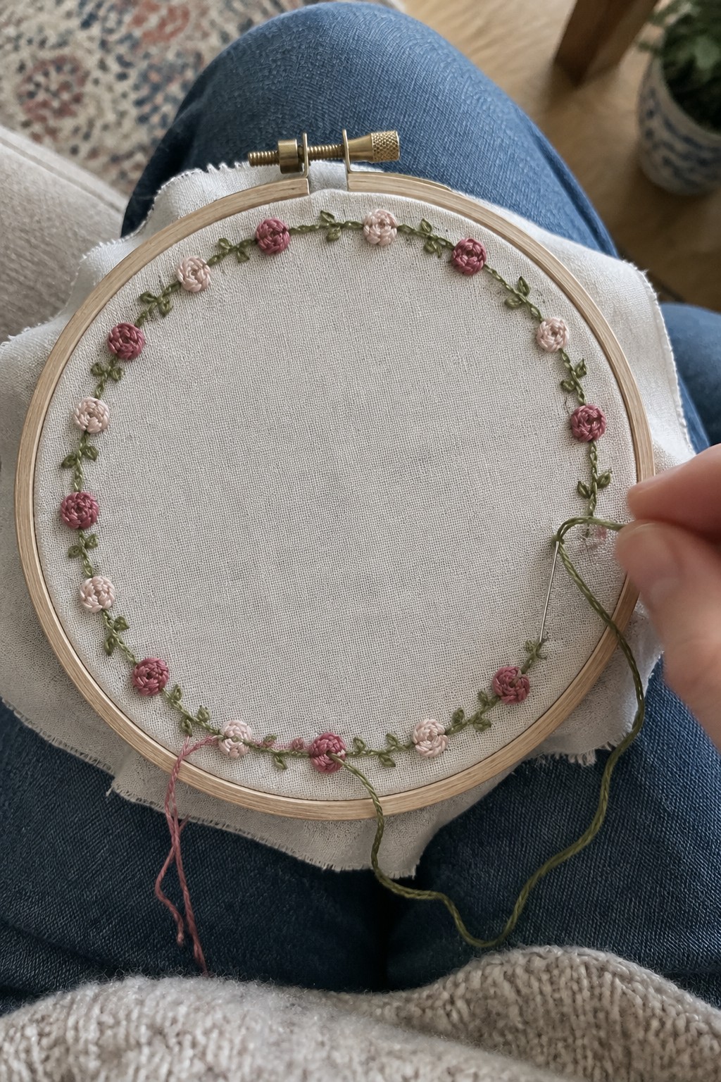 A circular vine border with alternating pink and pale blush flowers is being embroidered in a fabric hoop.