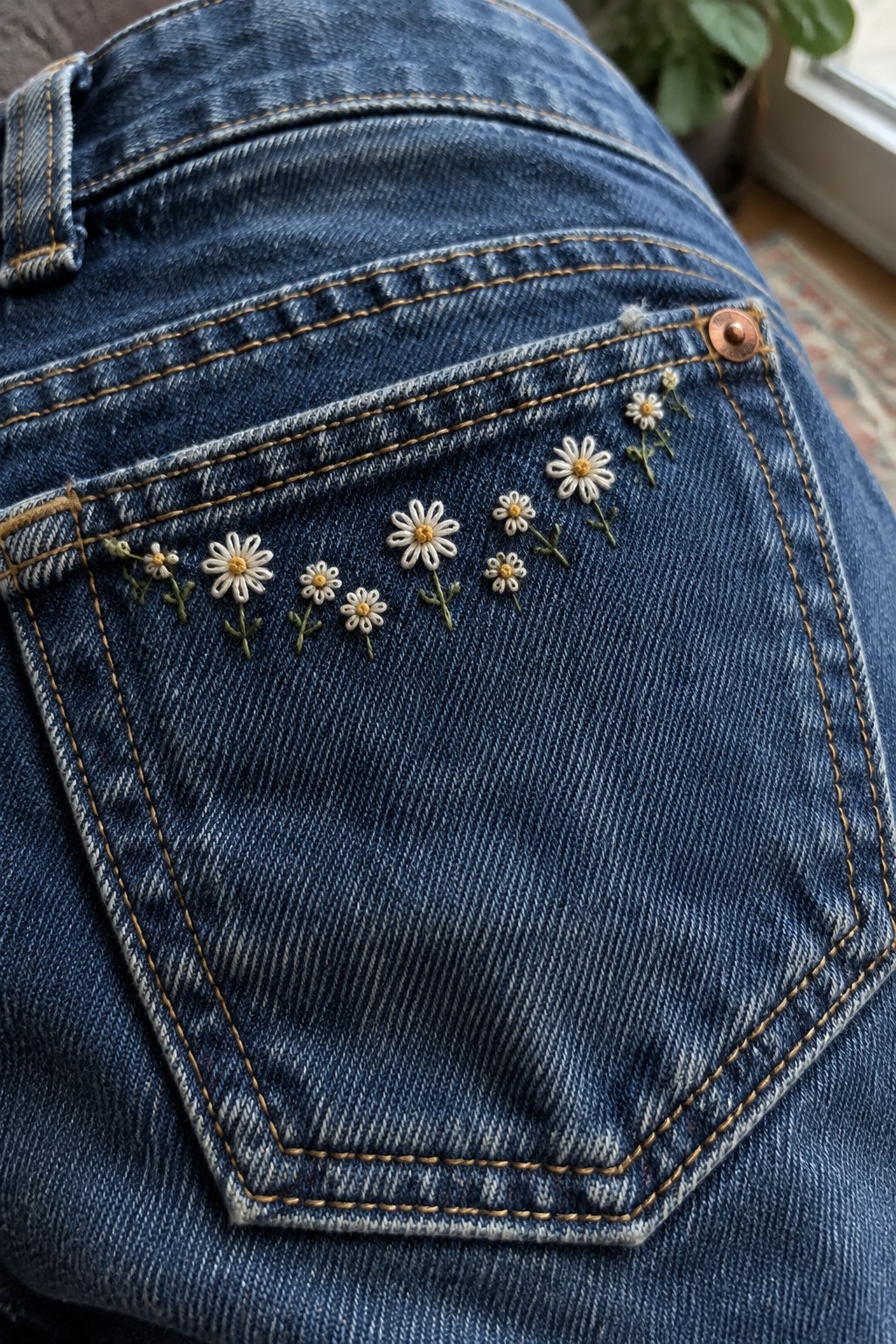 Cream and yellow embroidered daisies with green stems form a line across the top of a blue denim pocket.