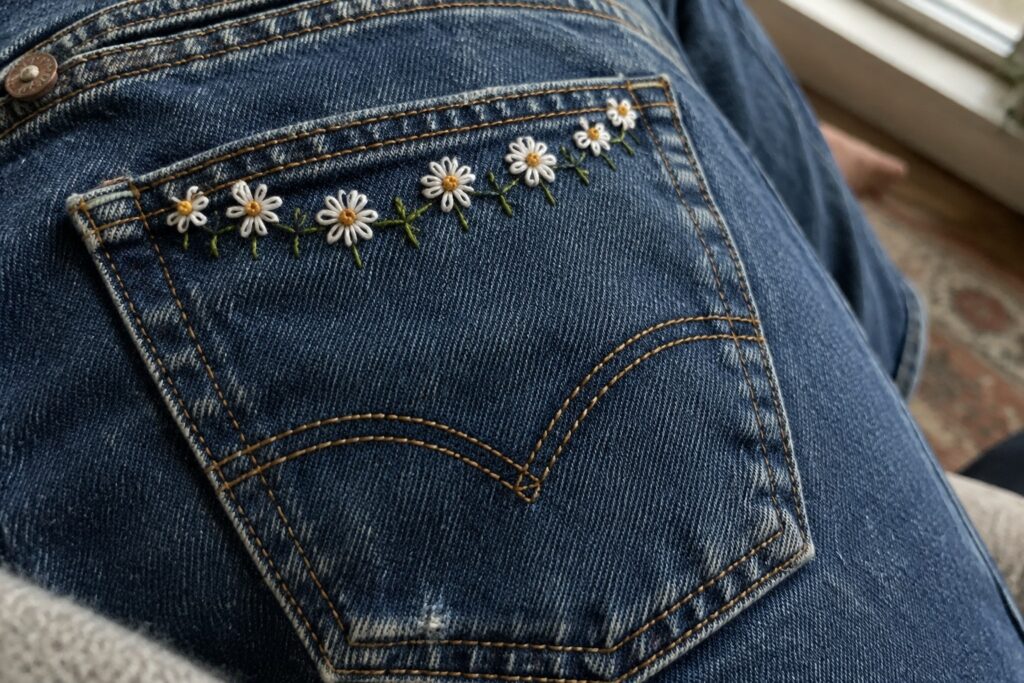 18 Charming Denim Pocket Embroidery Designs With Miniature Flowers Cream and yellow embroidered daisies with green stems form a line across the top of a blue denim pocket.