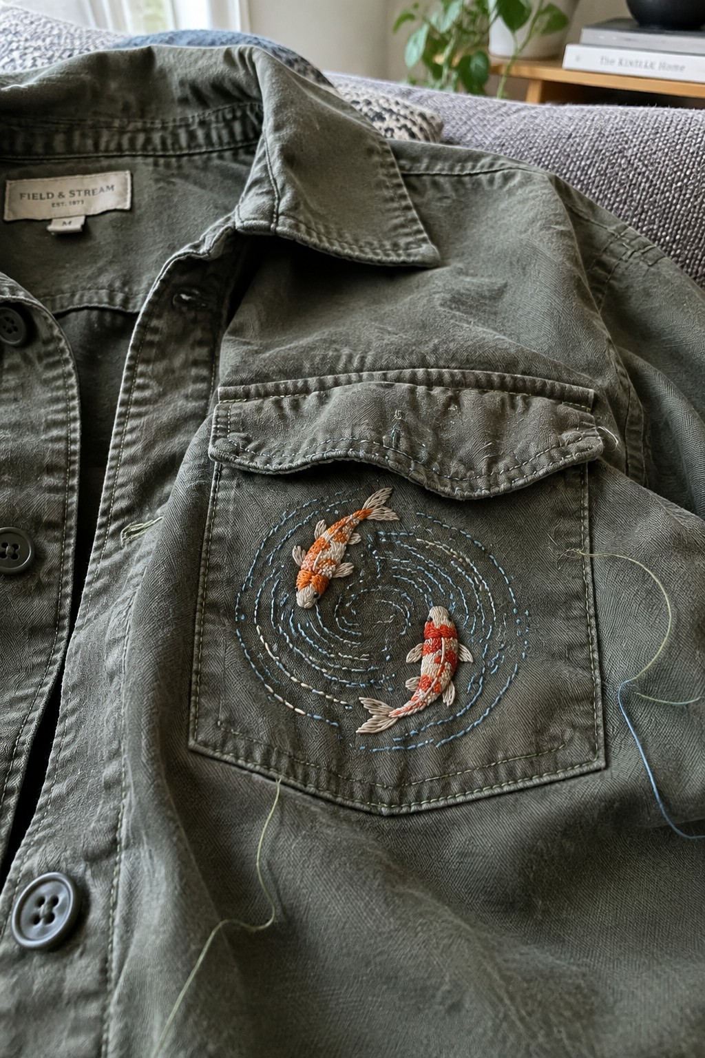 Two embroidered koi circle a blue stitched spiral on the pocket of an olive denim jacket.