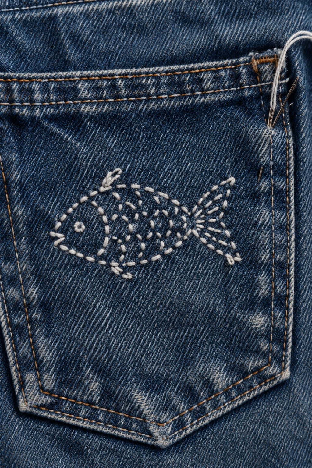 White stitched motif on the pocket of blue denim jeans, with a needle and thread near the top right.