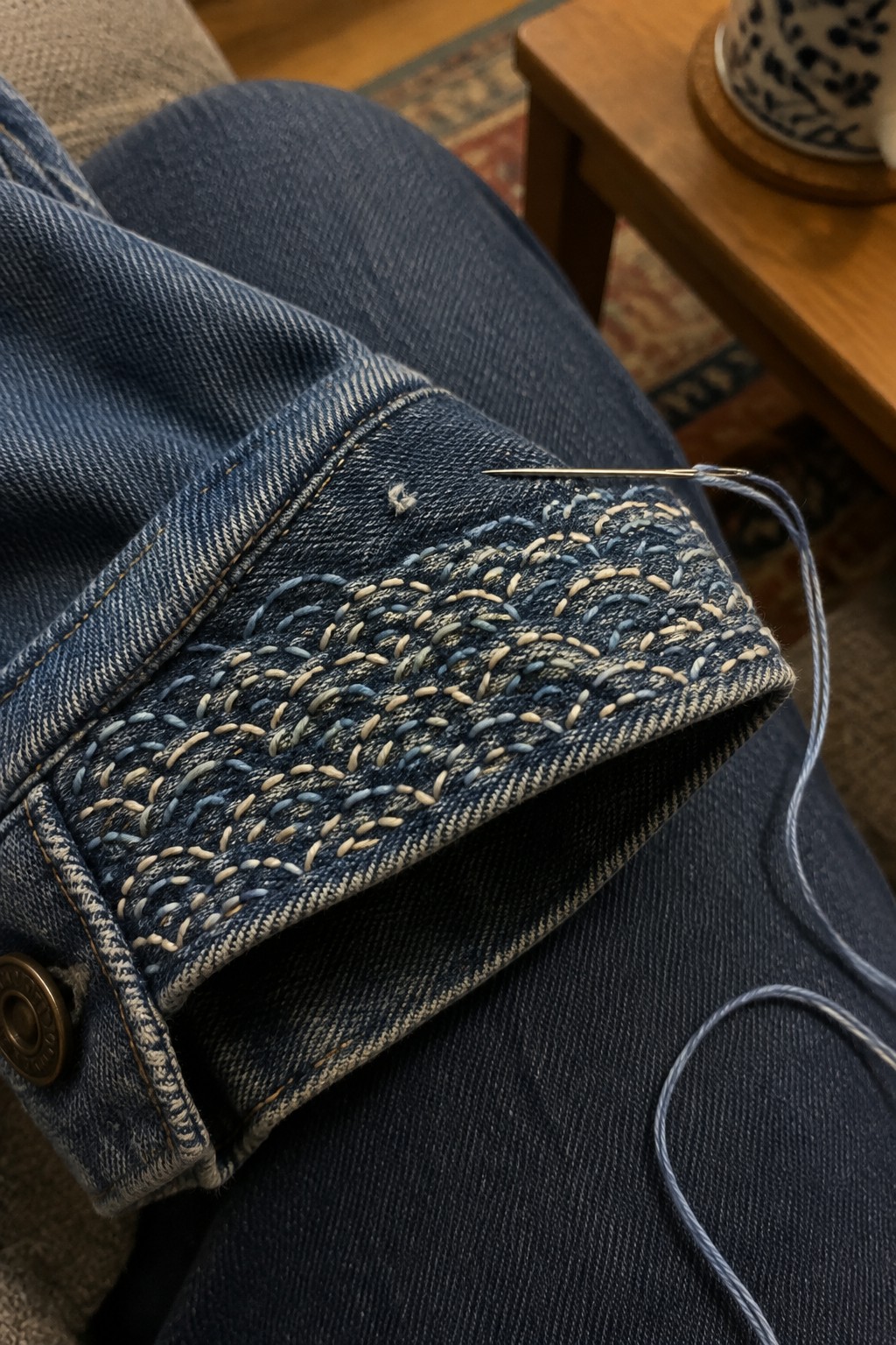 A jeans pocket is covered with dense cream and pale blue stitches, with a needle and thread resting above it.