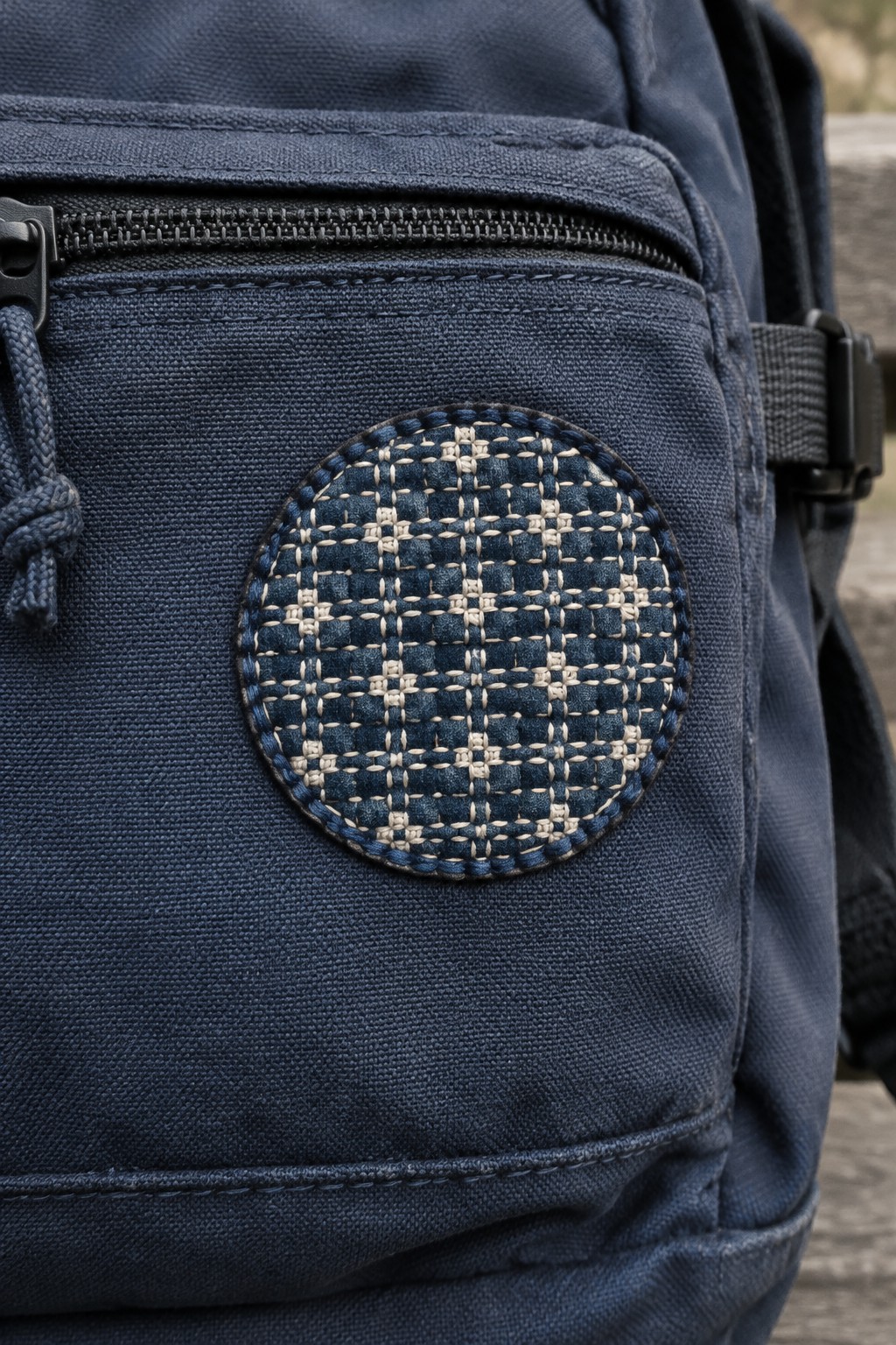 A circular cream-and-blue geometric stitched patch on the front pocket of a navy backpack.