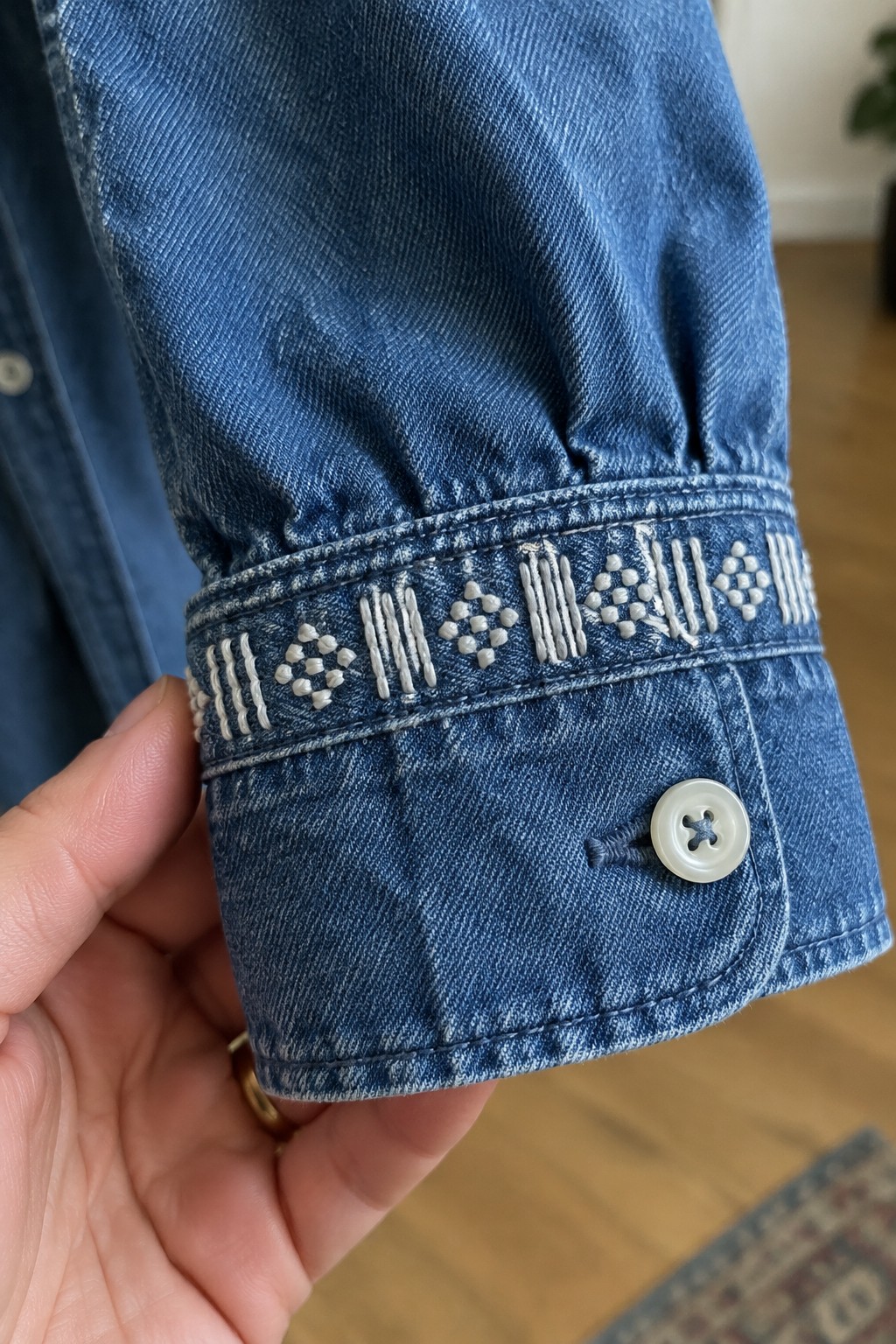 White geometric embroidery decorates the cuff of a blue denim shirt beside a button.