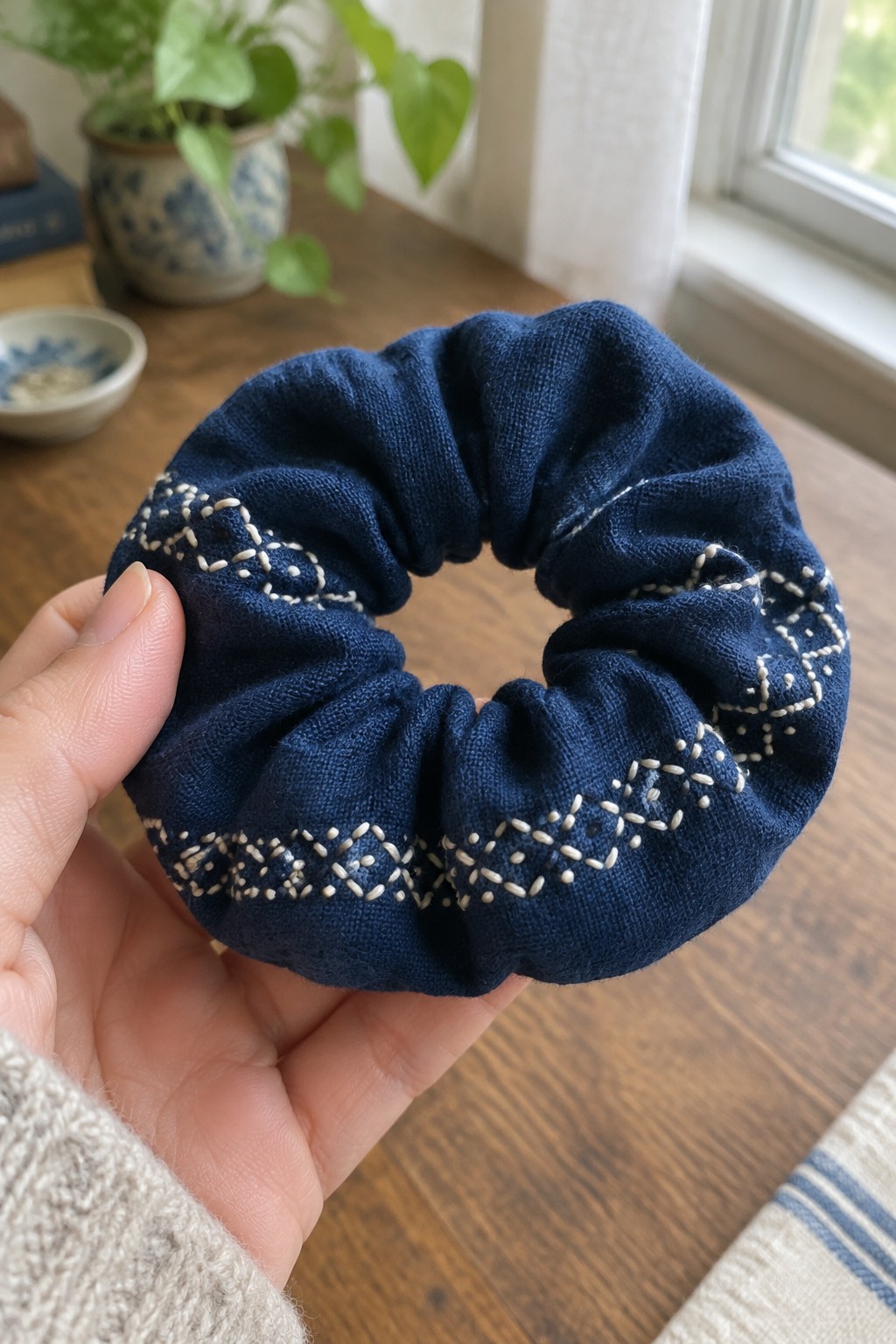 A hand holds a navy fabric scrunchie decorated with cream geometric embroidery.