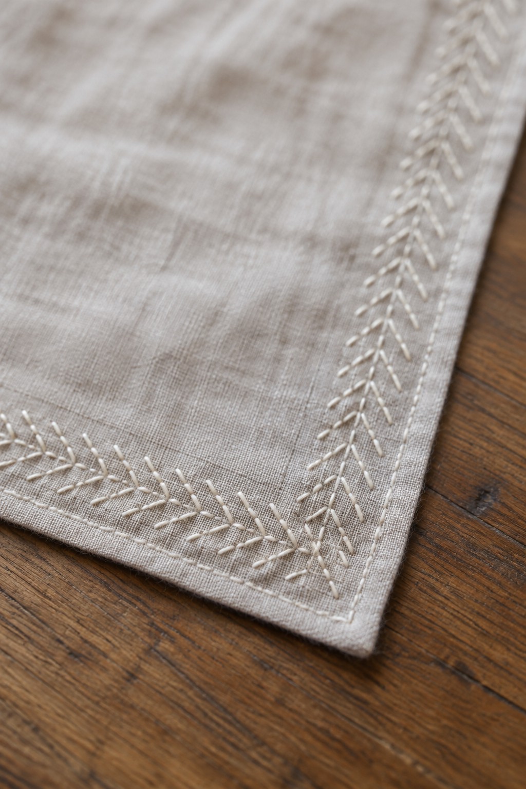 White chevron embroidery follows the corner edges of a beige linen napkin resting on a wooden table.
