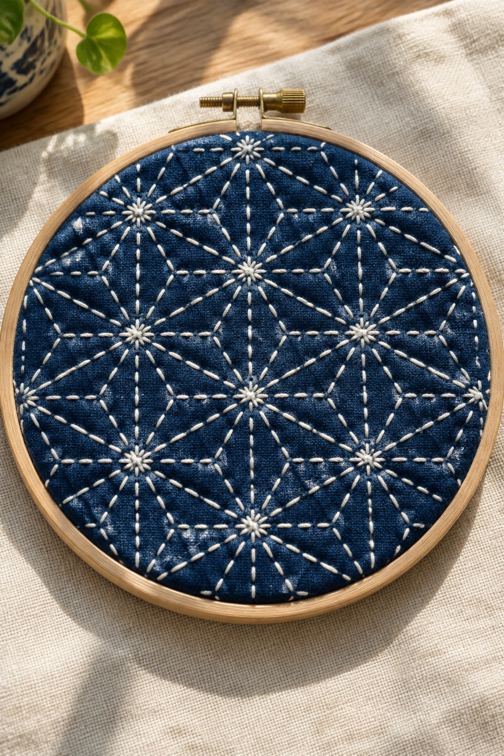 White geometric starburst stitches form a repeating pattern on indigo fabric held in a wooden embroidery hoop.
