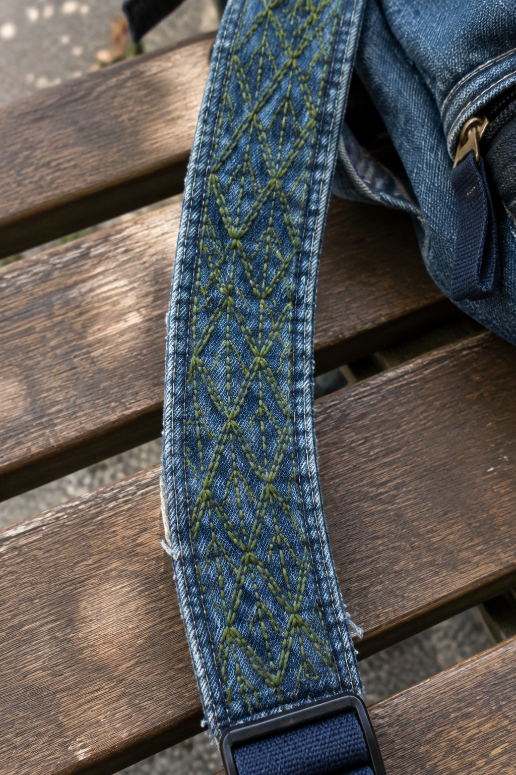 A denim bag strap with repeating green geometric stitching rests across a wooden bench.