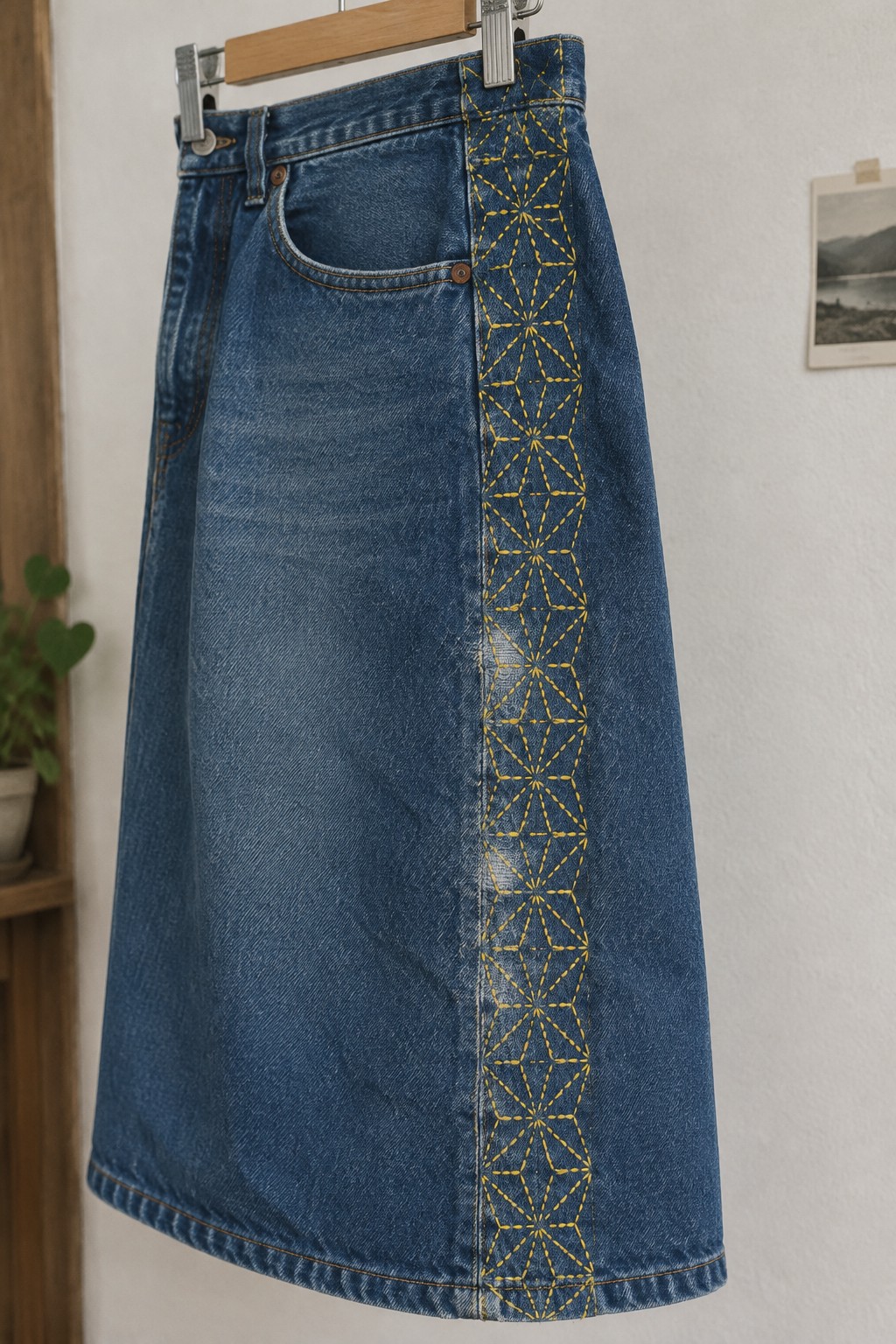 A blue denim skirt with a vertical panel of yellow geometric sashiko stitching hanging from a wooden hanger.