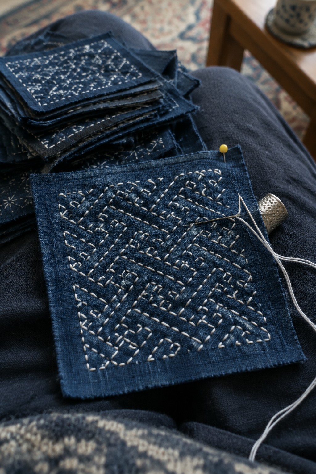 Cream geometric stitching covers an indigo fabric square beside a needle, thread, and stacked fabric blocks.