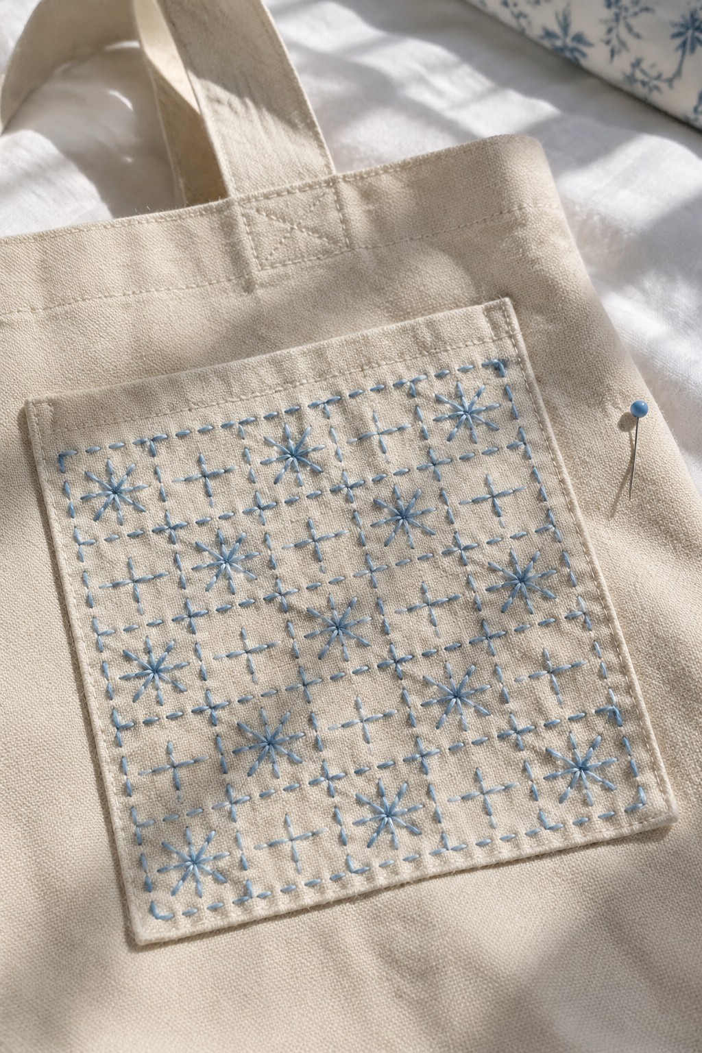 A beige tote bag with a pocket embroidered in blue geometric crosses, starbursts, and dashed lines.