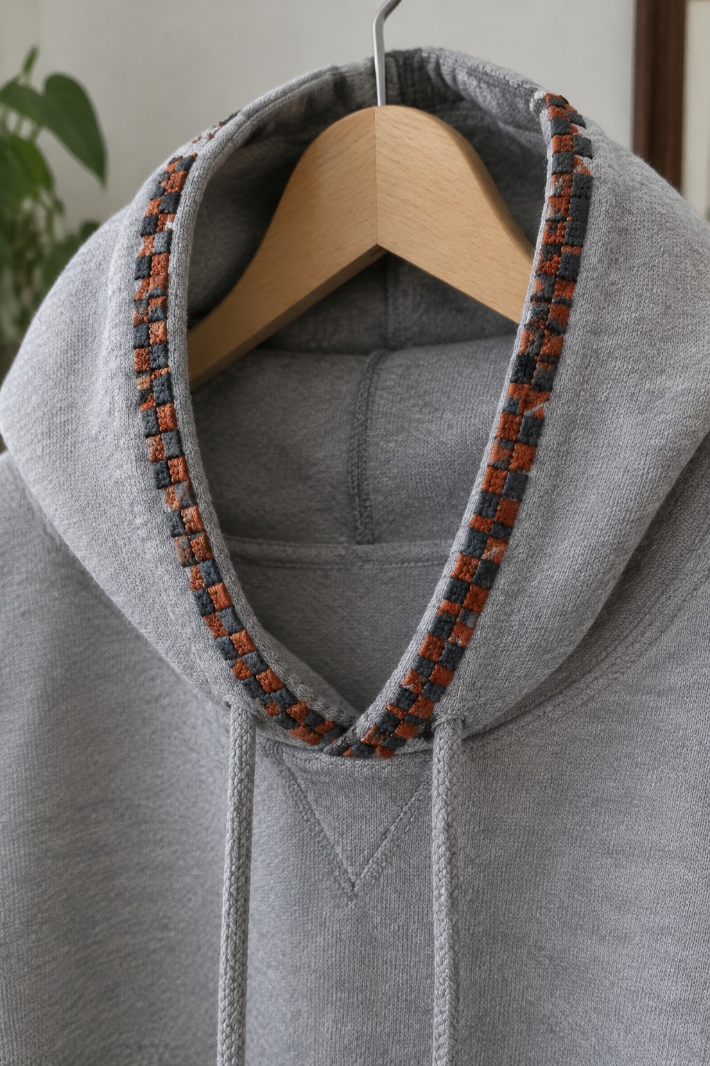 Gray hooded sweatshirt on a wooden hanger with rust and charcoal checkerboard embroidery along the hood edge.