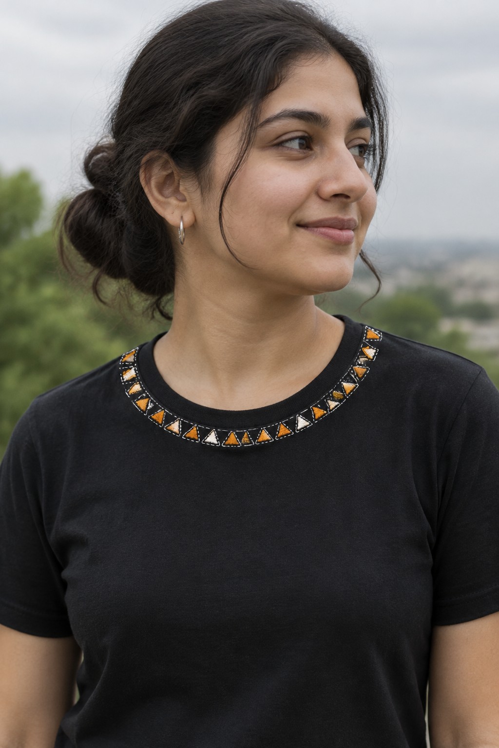 A woman wears a black T-shirt with a curved border of orange, cream, and dark triangle embroidery around the neckline.