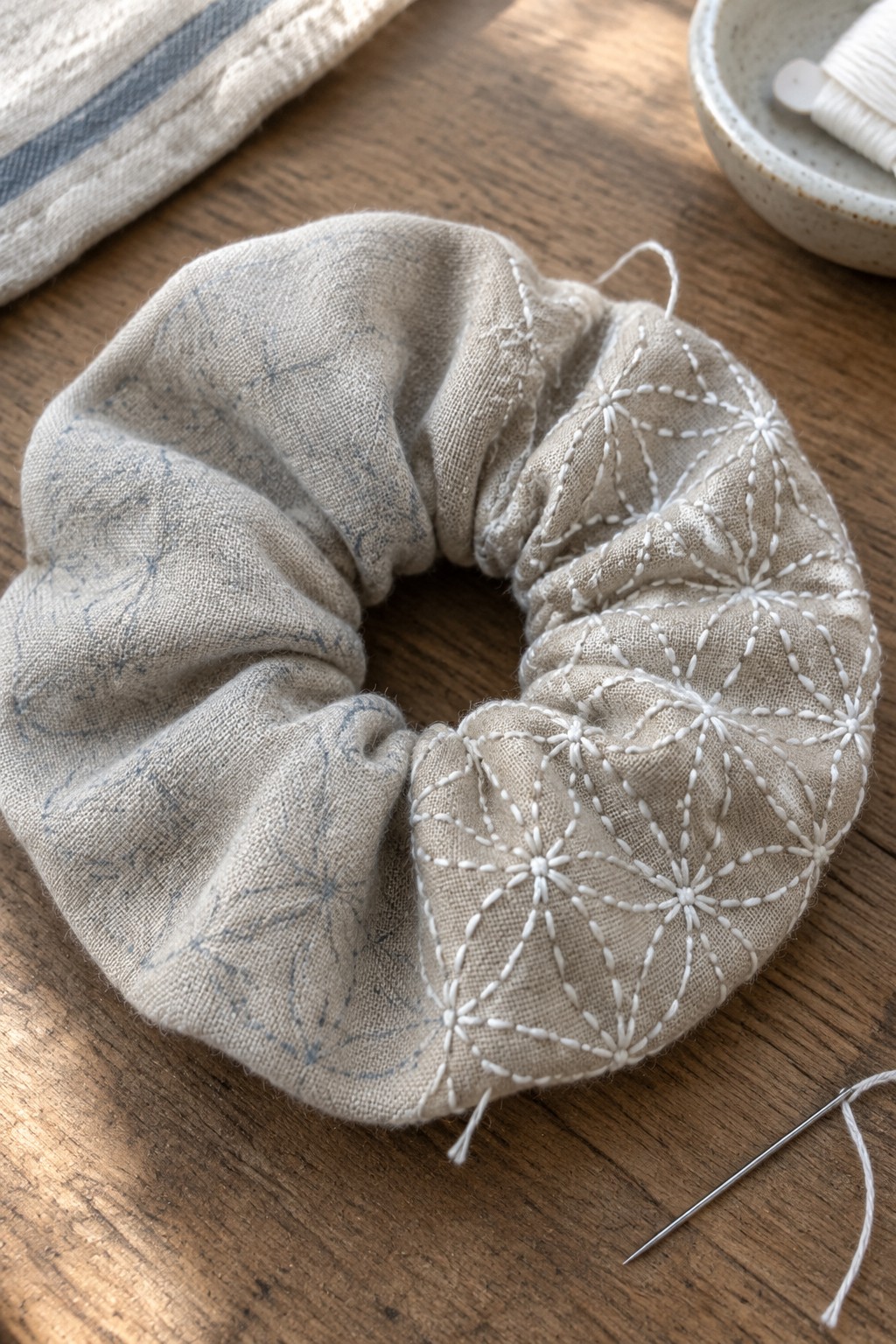 A gathered beige fabric scrunchie with white stitched patterns rests on a wooden table beside a needle and thread.