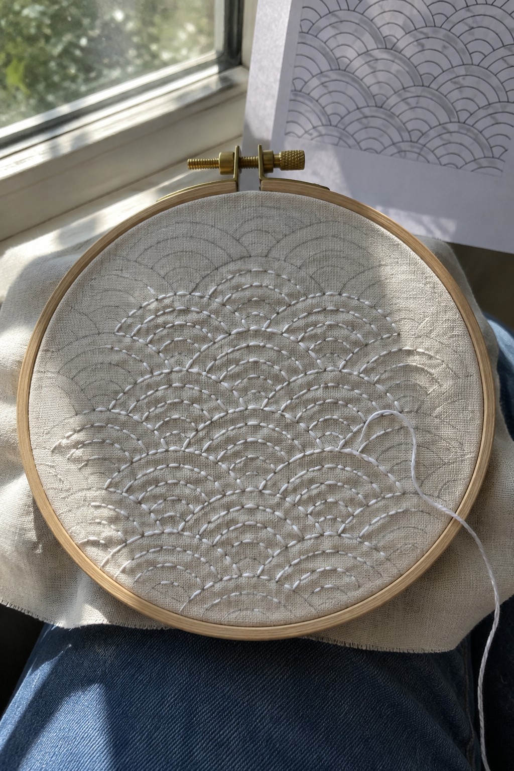 White curved stitches form repeating wave rows on fabric held in an embroidery hoop, with a matching pattern print nearby.