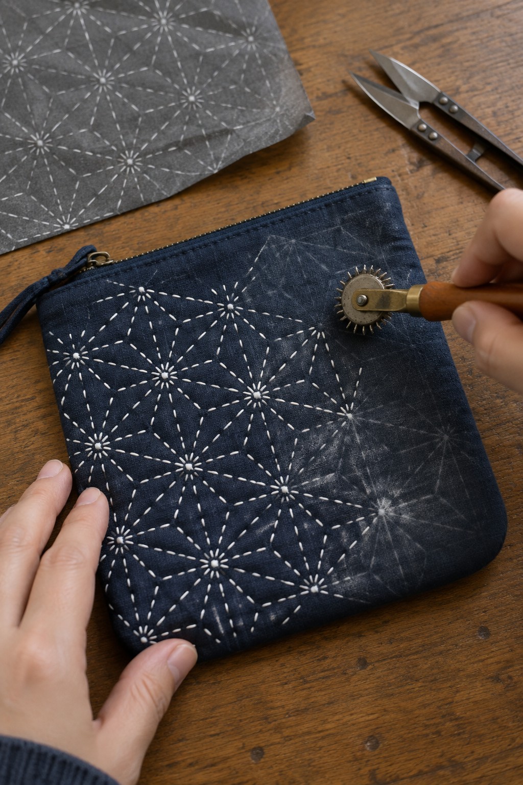White geometric sashiko stars are stitched across a navy zip pouch as a hand guides a tracing wheel over faint pattern marks.
