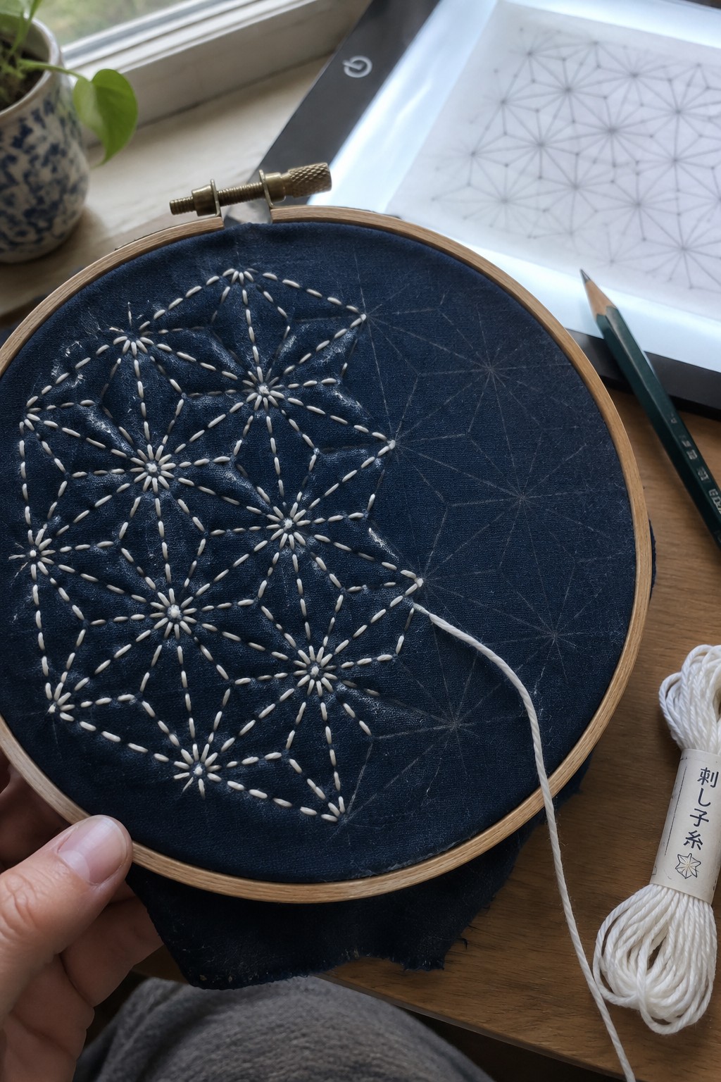 White geometric sashiko stitching covers part of dark blue fabric held in an embroidery hoop, with the pattern traced on the unstitched side.