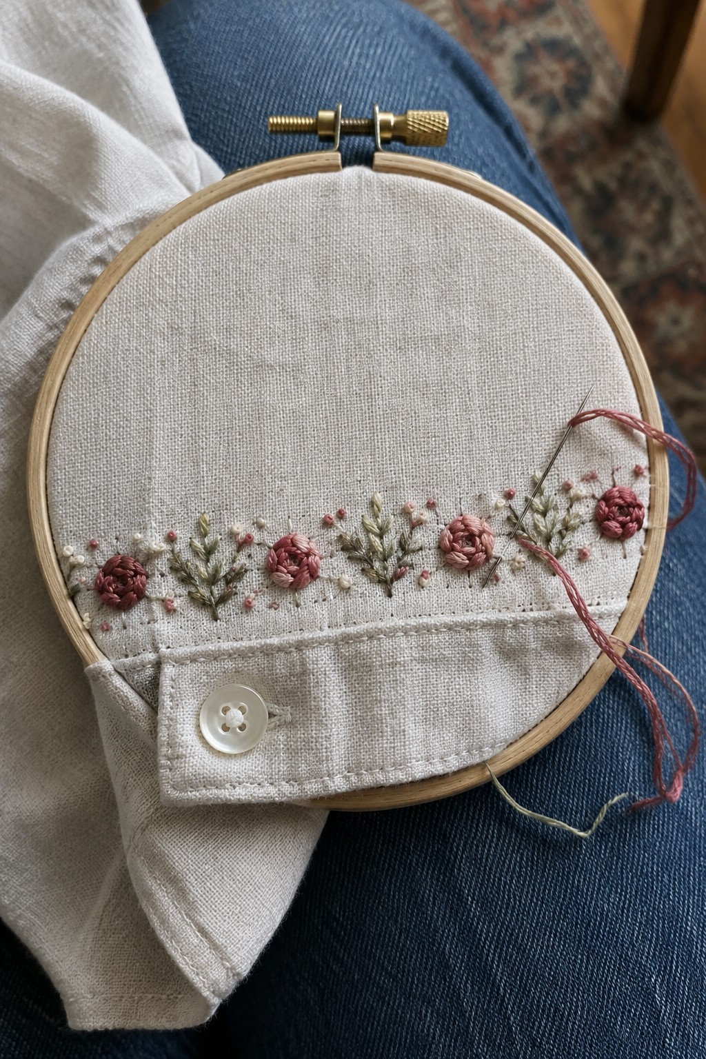 A hoop holds a linen cuff embroidered with small rose-colored flowers and leafy sprigs.