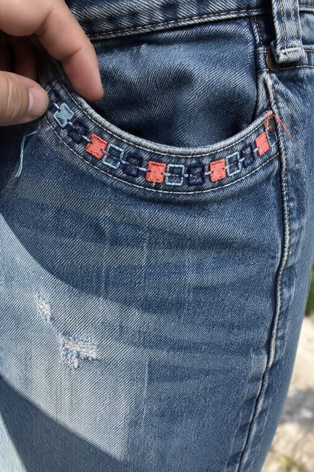 Coral, pale blue, and dark blue geometric embroidery follows the curved edge of a jeans pocket.