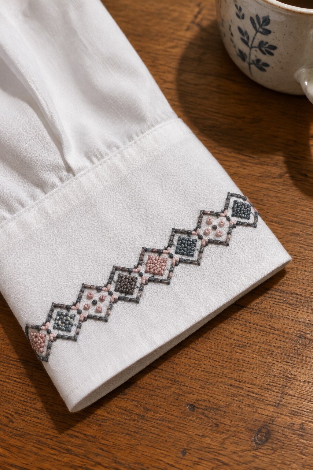 A white shirt cuff embroidered with a stepped border of interlocking dark gray and blush geometric shapes.