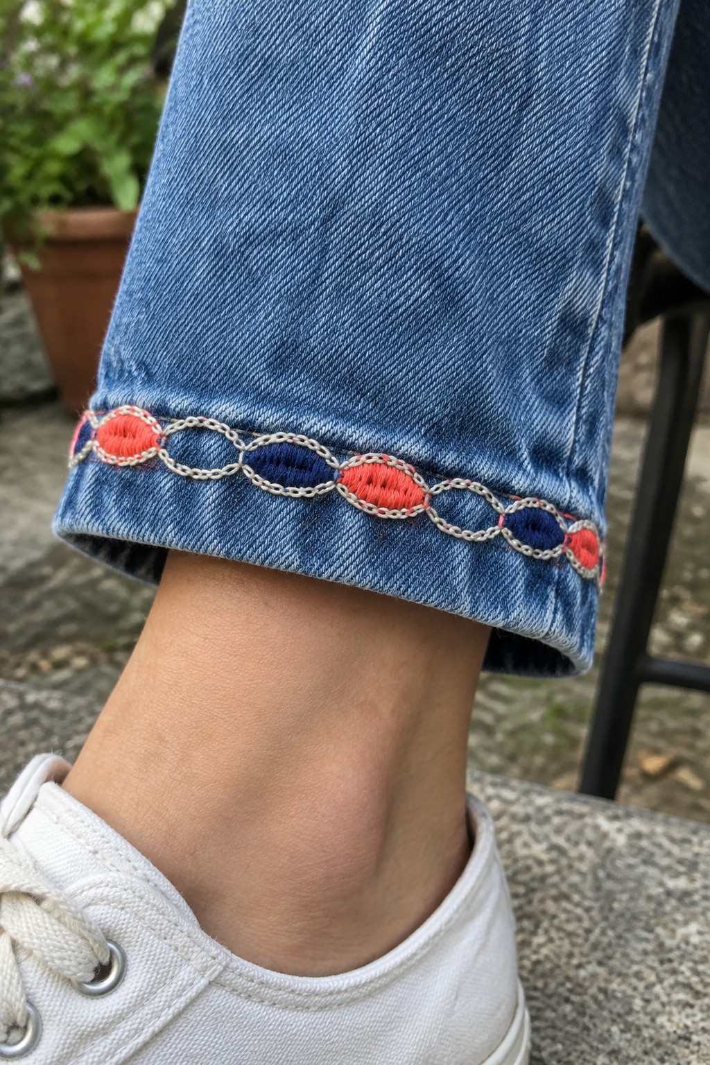 Coral and navy interlocking oval embroidery decorates the turned-up cuff of blue jeans above a white sneaker.