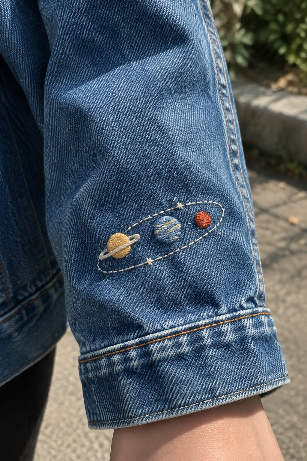 Three embroidered planets and small stars sit inside a dashed orbit on a denim jacket sleeve.