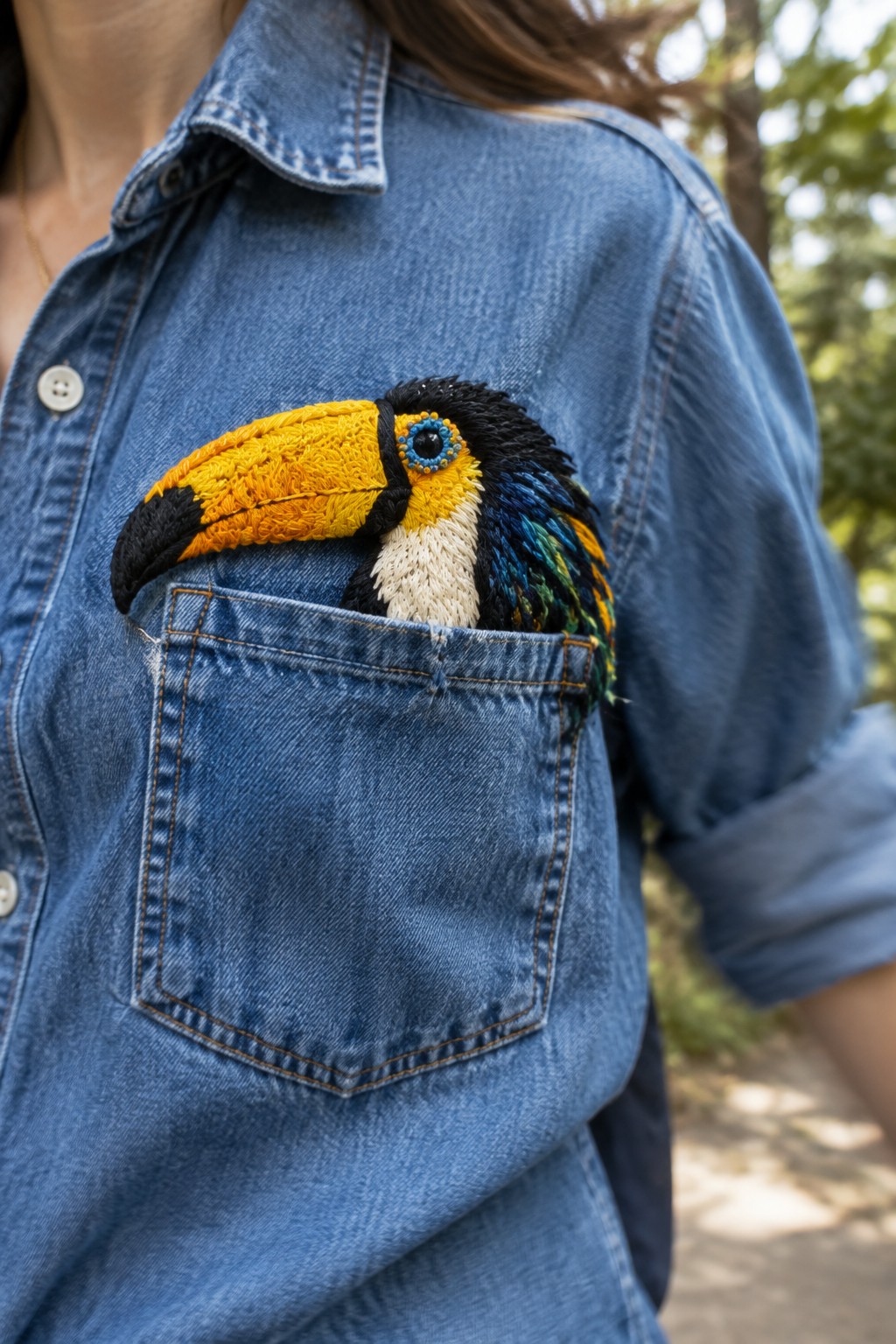 A colorful embroidered toucan peeks out of the chest pocket of a blue denim shirt.