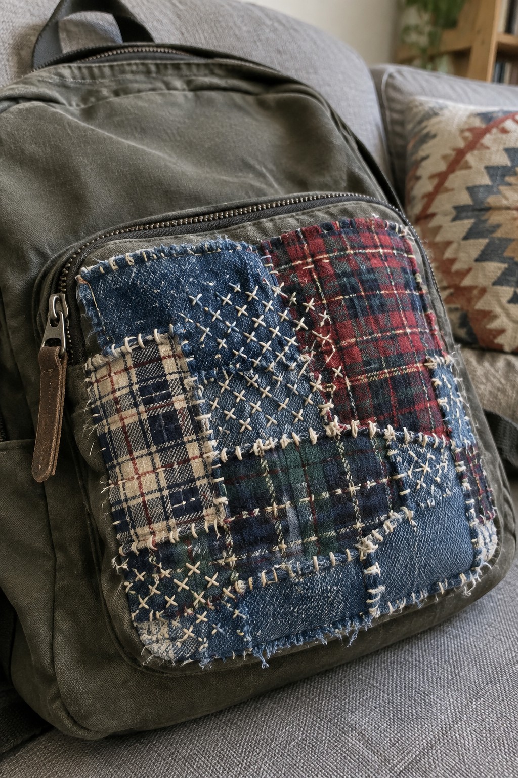 An olive backpack with layered plaid and denim patches, secured with visible cream stitching across the front pocket.