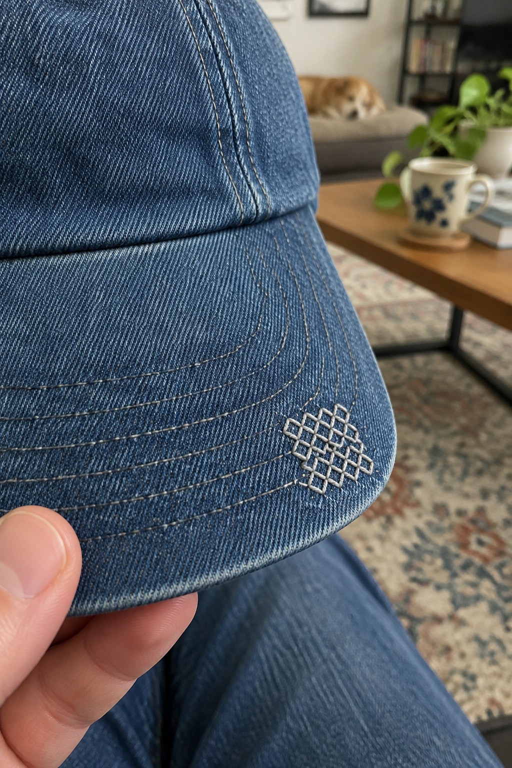 A denim cap brim with a small pale crosshatched embroidery motif beside curved rows of stitching.