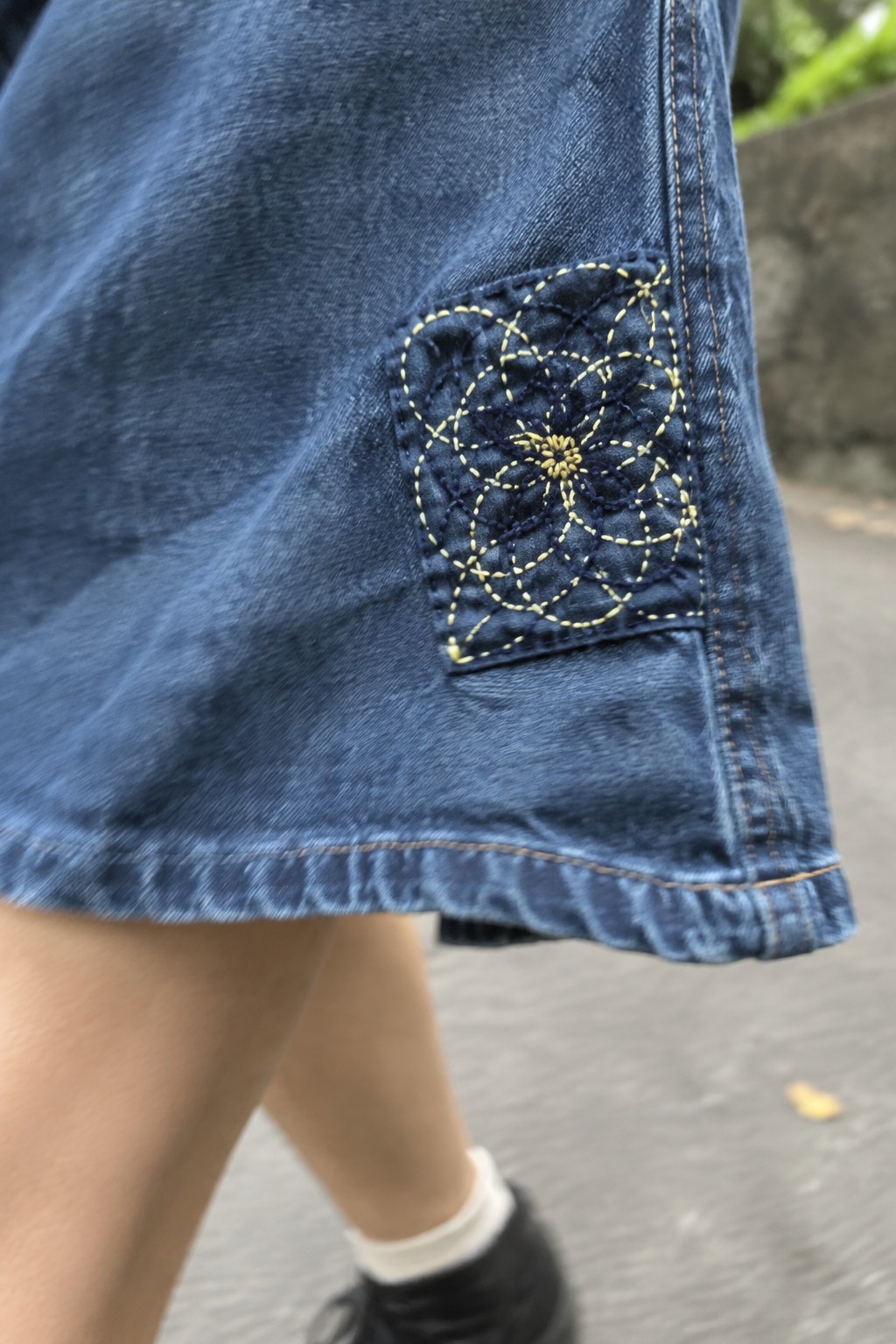 A dark square patch with pale yellow interlocking flower stitches is attached near the hem of blue denim.