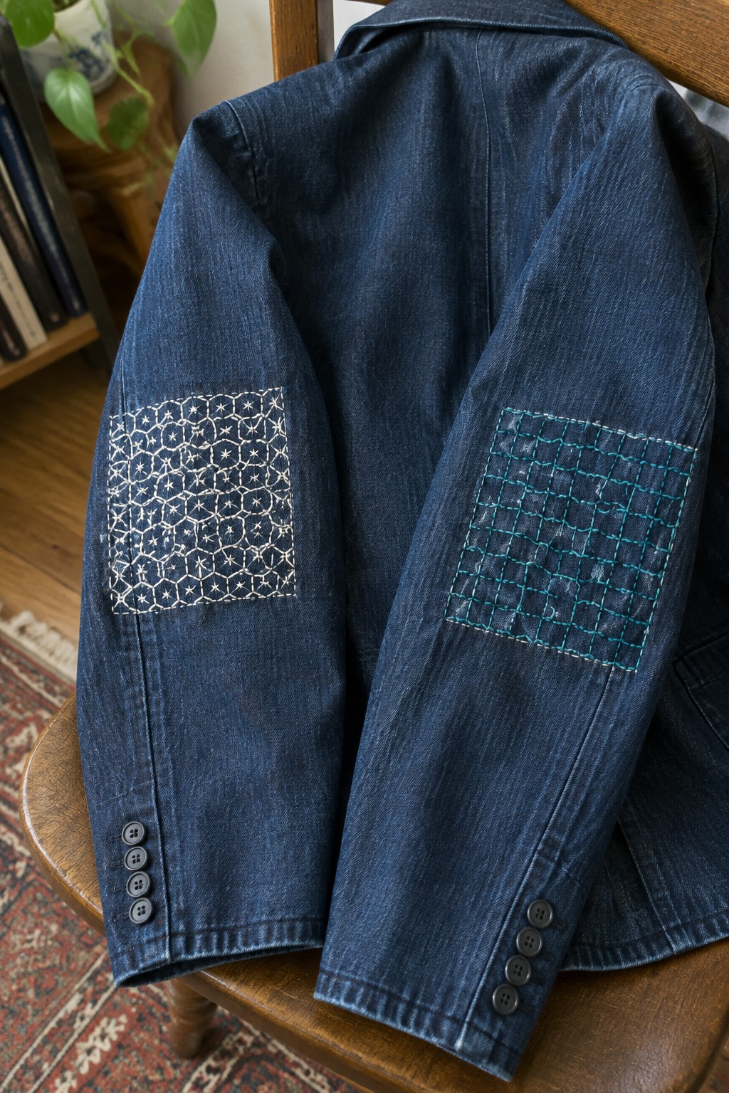 A dark denim jacket draped over a wooden chair has a white geometric embroidered square on one sleeve and a turquoise grid patch on the other.
