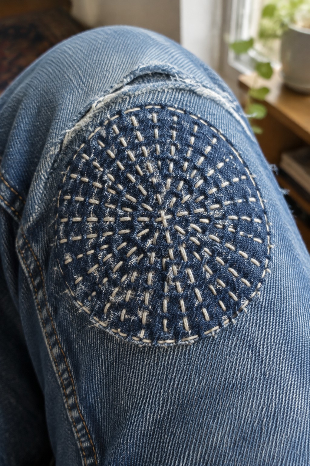 A dark circular patch with radiating white stitches covers the knee of blue jeans.
